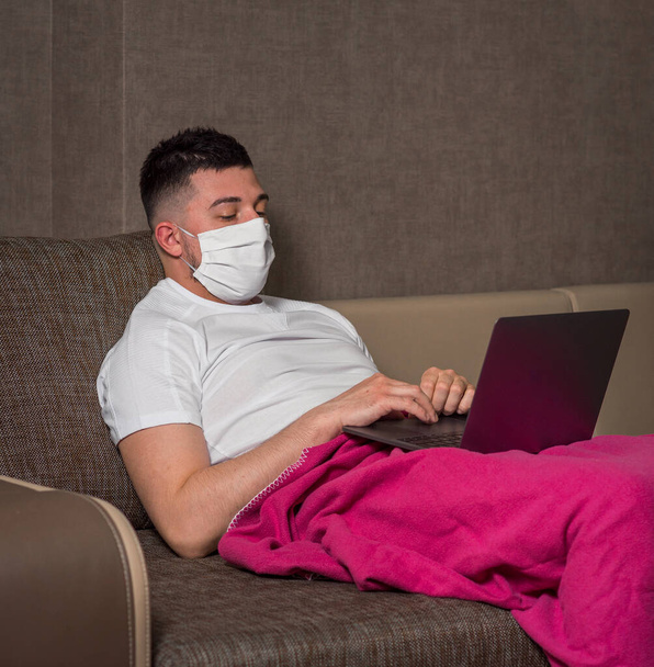 A white man in a medical mask works from home during infection with a coronavirus or Covid-19, a patient in a quarantine ward using a computer. legs under the covers. home insulation - 写真・画像