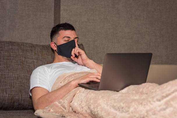 Un hombre blanco con una máscara médica trabaja desde casa durante la infección con un coronavirus o Covid-19, un paciente en una sala de cuarentena usando una computadora. piernas debajo de las sábanas. aislamiento del hogar
 - Foto, imagen