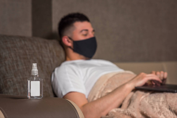 Fertőtlenítő a háttérben. egy férfi orvosi maszkban otthon dolgozik egy koronavírus vagy Covid-19 fertőzés alatt számítógéppel. lábak a takaró alatt. otthoni szigetelés - Fotó, kép