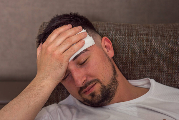 man is sick in bed at home. headache. white rag on the head. bandaged head of a bearded man. coronavirus, covid 19, quarantine - Photo, image