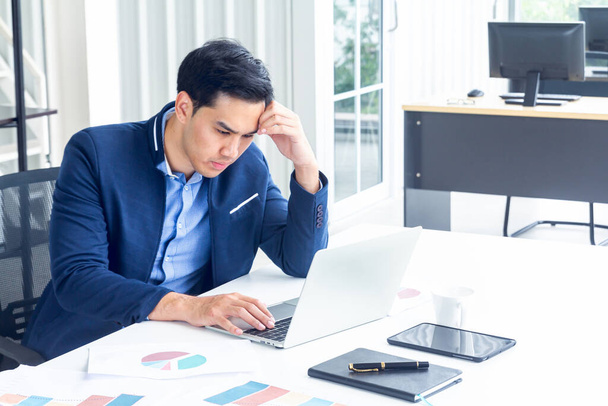 A young businessman sitting in a modern office. He has a feel stressed about the result of business profits not positive. On his table have a computer laptop tablet pen paper graph and a coffee cup. - Foto, Bild