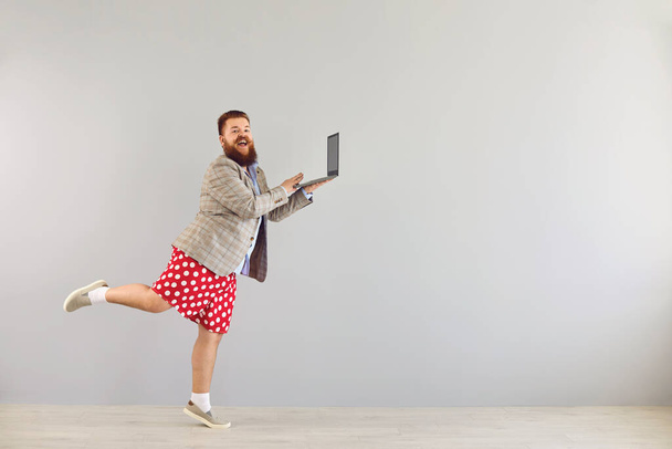 Vtipný tlustý muž v bundě pracuje pomocí notebooku při tanci na šedém pozadí. - Fotografie, Obrázek