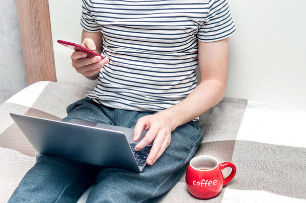 Young man working on a laptop in bed at home and big red cup beside.A man looks at a smartphone. Concept Working remotely - Valokuva, kuva