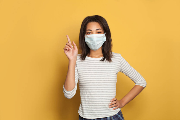 Junge Afroamerikanerin mit medizinischer Gesichtsmaske zeigt mit dem Finger nach oben auf gelbem Studiohintergrund - Foto, Bild
