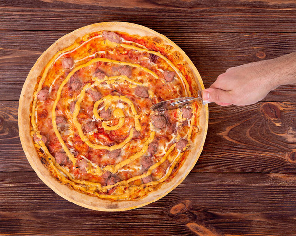 Man's hand cuts the salsiccia pizza with a pizza cutter, top view. Delicious pizza with beef sausages, mozzarella, various sauces and marinated red onions on round wood platter which is on wooden rustic table - Photo, Image