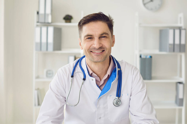 Male doctor with a stethoscope smiling while sitting at a table in a white interior of a clinic office. - Foto, imagen
