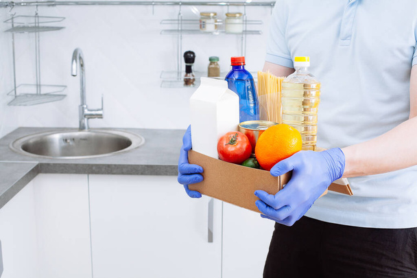 Courier in protective medical gloves delivers box with set of fresh raw vegetables and  food supplies crisis food stock for quarantine isolation period in a modern kitchen room background. Organic food and products delivery - Φωτογραφία, εικόνα