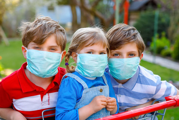 tři děti, malá batolata a dva chlapci v lékařské masce jako ochranu proti pandemické koronavirové chorobě. Děti s nákupním vozíkem pomocí ochranných pomůcek pro boj proti covid 19. - Fotografie, Obrázek