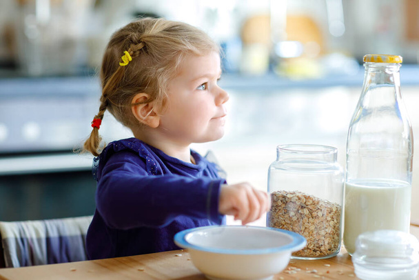 Adorable toddler girl eating healthy oatmeals with milk for breakfast. Cute happy baby child in colorful clothes sitting in kitchen and having fun with preparing oats, cereals. Indoors at home - Photo, Image