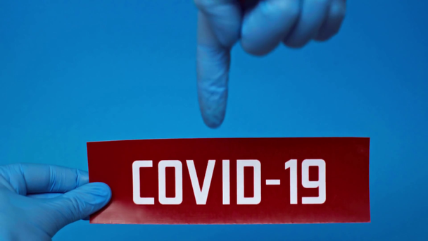 przycięty widok człowieka w lateksowych rękawiczkach wskazujących na kartę covid-19 odizolowaną na niebiesko - Materiał filmowy, wideo