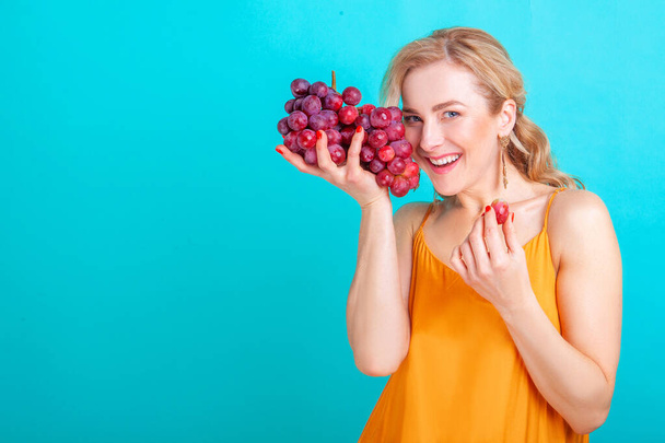 blond vrouw holding een bos van druiven op een blauwe achtergrond in de studio - Foto, afbeelding