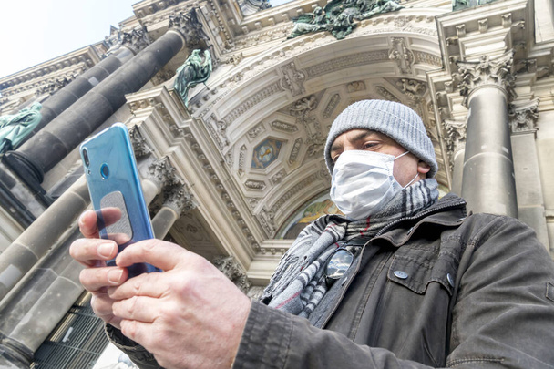 Turismo durante brote de enfermedad, pandemia del Coronavirus COVID-19 SARS-CoV-2. Hombre cubriéndose la cara con una máscara médica para protegerse la nariz y la boca usando su teléfono inteligente fuera de la Catedral de Berlín
 - Foto, imagen