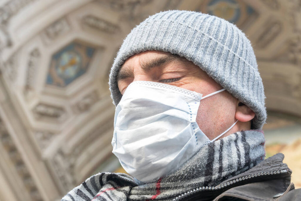 Epidemie onemocnění, pandemie COVID-19 (Coronavirus SARS-CoV-2). Snímek hlavy muže se zavřenýma očima, který si zakrývá tvář lékařskou maskou, aby si chránil nos a ústa venku. - Fotografie, Obrázek