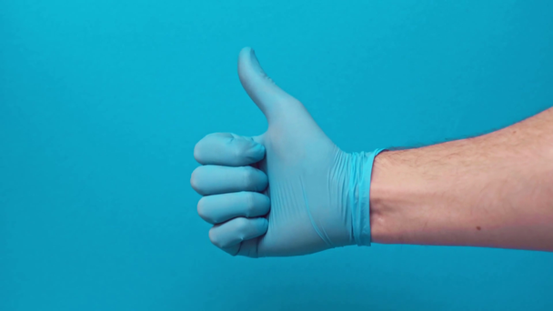 ανδρικό χέρι σε λαστιχένιο γάντι με τον αντίχειρα επάνω απομονωμένο σε μπλε, covid-19 έννοια - Πλάνα, βίντεο