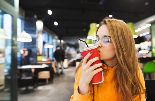 Jolie fille en vêtements décontractés dans des lunettes se trouve dans un restaurant de restauration rapide et embrasse un verre rouge avec du soda. Dame aux cheveux blonds embrasse un verre de cola sur fond de café. Espace de copie
 - Photo, image