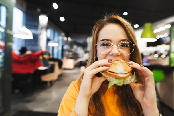 Bella ragazza in occhiali e capelli biondi mangiare un hamburger appetitoso e guardando nella fotocamera con un volto serio.Lady morde un grande hamburger sullo sfondo di un ristorante fast food. Pranzo fast food
 - Foto, immagini