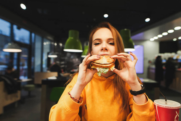 Chica atractiva en un suéter naranja se come una hamburguesa en un café, mira a la cámara. Bonita estudiante comiendo comida chatarra en restaurante de comida rápida sosteniendo hamburguesa con queso en la mano
. - Foto, imagen