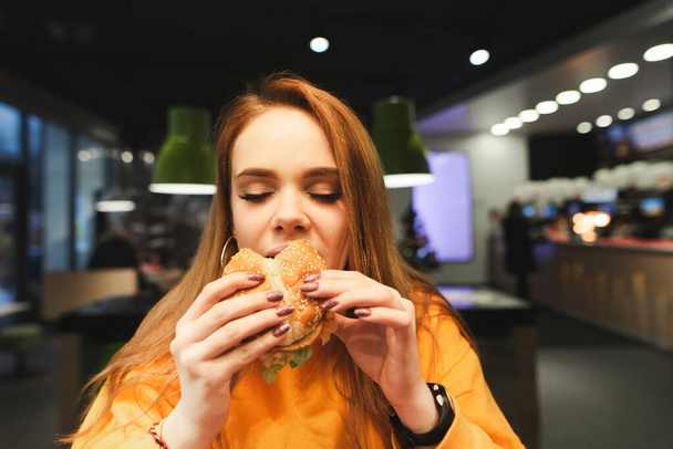 Szoros portré egy csinos hölgyről, aki csukott szemmel hamburgert harap és jól érzi magát. Csinos lány eszik ízletes junk food a háttérben elmosódott gyorsétterem. A hölgy gyorskaját eszik.. - Fotó, kép