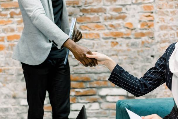 Business concept of hand shaking. Cropped image of shaking hands after agreement between business lady in black jacket and dark-skinned businessman in the modern loft office room - Фото, зображення