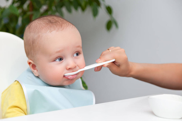 Little baby boy in color clothes eating yoghurt from spoon, dirty mouth - Photo, Image