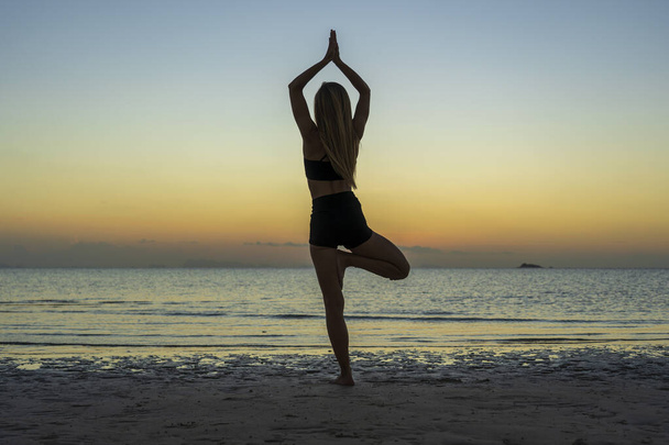 Silhouette of woman standing at yoga pose on the tropical beach during sunset. Caucasian girl practicing yoga near sea water - Foto, Bild