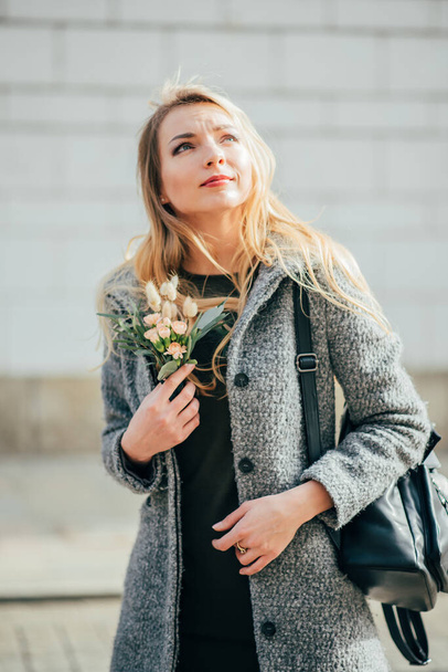 Caucasian beautiful young  blonde woman in grey coat is walking in Krakow city streets. young pretty blond woman with long blond wavy hair smiles to people in the street and holding her blond hair, the backpack and little bouquet. - Фото, изображение
