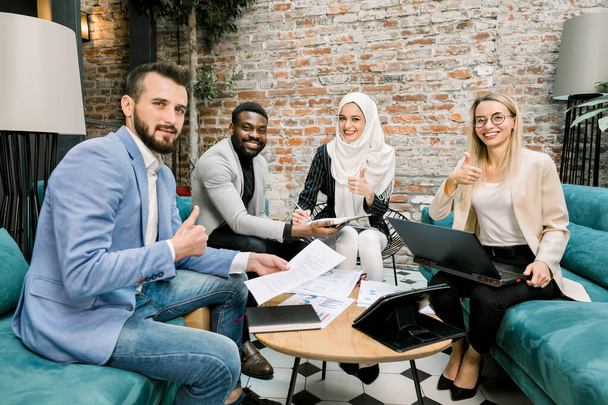 Successful excited multi-ethnic group of business people sitting together in office around the table with papers and laptop, showing thumbs up gesture, looking at camera and smiling - Zdjęcie, obraz