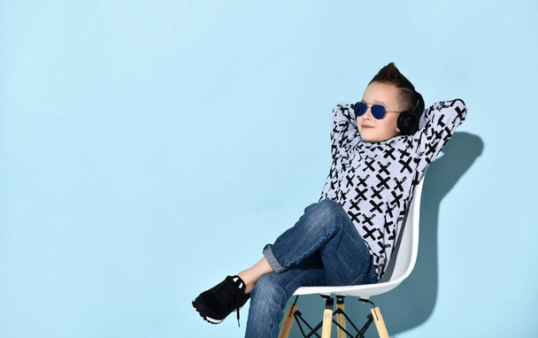Teenage child in headphones, sunglasses, blue jeans, hoodie, sneakers. Propping chin by fist, sitting on chair. Blue background - Photo, image