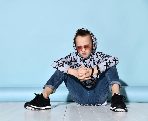 Teenager in sunglasses, blue jeans, hoodie, black bracelet and sneakers. Raised hand up, sitting on white floor. Blue background - 写真・画像
