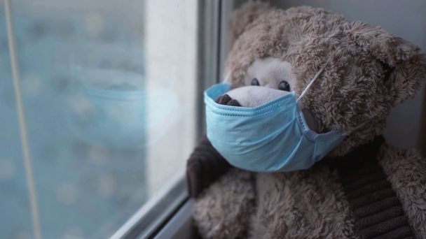 αρκουδάκι σε μπλε ιατρική μάσκα κοντά στο παράθυρο  - Πλάνα, βίντεο