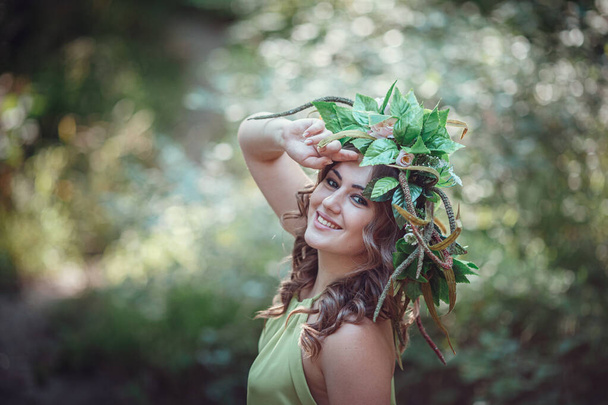 Mooie jonge vrouw in een groene jurk en een krans in een zonnig bos. ceremonie op midzomer- of aardedag. idee en concept van parfum, sprookjes en geluk - Foto, afbeelding