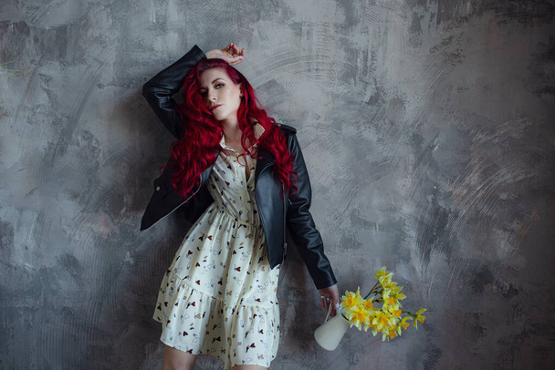 young beautiful red haired woman holding bouquet of fresh yellow daffodils, isolated on grey wall background. The girl is wearing a dress, a leather jacket and yellow shoes - Photo, Image