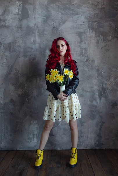 young beautiful red haired woman holding bouquet of fresh yellow daffodils, isolated on grey wall background. The girl is wearing a dress, a leather jacket and yellow shoes - Fotoğraf, Görsel
