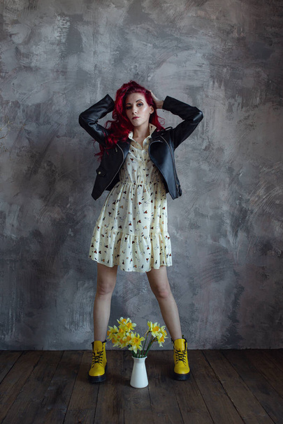 young beautiful red haired woman holding bouquet of fresh yellow daffodils, isolated on grey wall background. The girl is wearing a dress, a leather jacket and yellow shoes - Fotó, kép