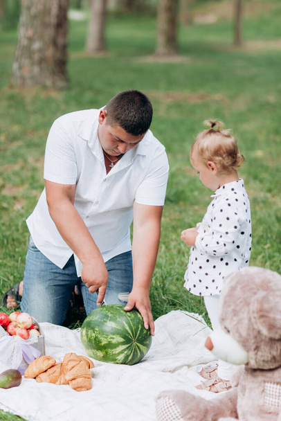 Tata i córeczka na pikniku w parku w letni dzień. Młody ojciec kroi arbuza. Dzień ojca, dziecka. Wesołych świąt - Zdjęcie, obraz