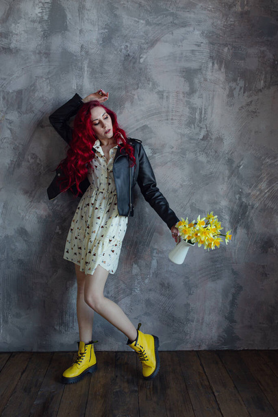 若い美しい赤い髪の女性は、灰色の壁の背景に隔離された新鮮な黄色の水仙の花束を持っています。女の子はドレス、革のジャケットと黄色の靴を着ている - 写真・画像