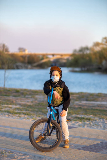Πορτρέτο ενός εφήβου στο ηλιοβασίλεμα με μάσκα σε μπλε ποδήλατο. Καραντίνα. Απαγορεύεται η οδήγηση ποδηλάτου. Covid-19. - Φωτογραφία, εικόνα