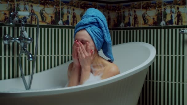 Young 30s woman washing face sitting in bathtub with foam at green bathroom at home. Woman with blue towel on head relaxing in bathtub. Home spa concept. Close up - Materiał filmowy, wideo
