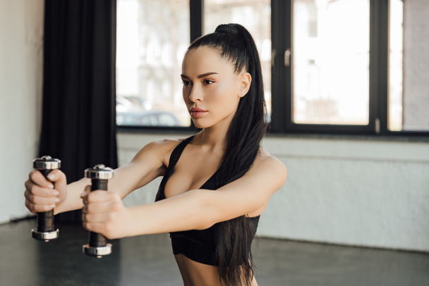 Beautiful sportswoman holding dumbbells with outstretched hands in gym - Valokuva, kuva