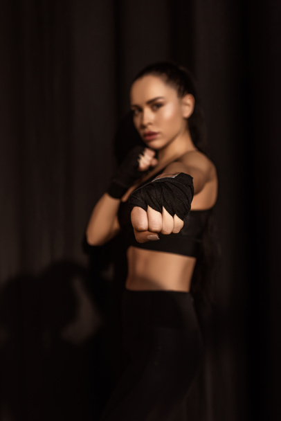 Selektiver Fokus der Sportlerin in Kampfposition, die die Faust mit ausgestreckter Hand zeigt und in die Kamera auf schwarz blickt - Foto, Bild