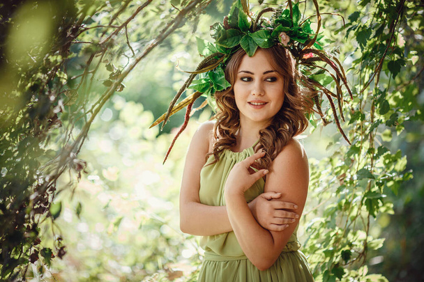 Beautiful young woman in a green dress and a wreath in a sunny forest. ceremony on Midsummer or Earth Day. idea and concept of perfume, fairy tales and happiness - Photo, Image