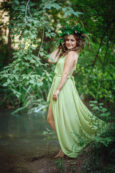 Όμορφη νεαρή γυναίκα με πράσινο φόρεμα και στεφάνι σε ένα ηλιόλουστο δάσος. τελετή στα μέσα του καλοκαιριού ή την Ημέρα της Γης. ιδέα και έννοια του αρώματος, παραμύθια και ευτυχία - Φωτογραφία, εικόνα
