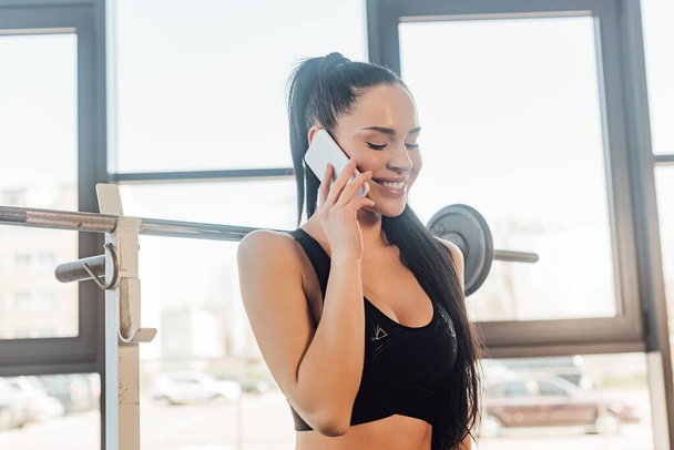 Beautiful sportswoman smiling and talking on smartphone near barbell in gym - Zdjęcie, obraz