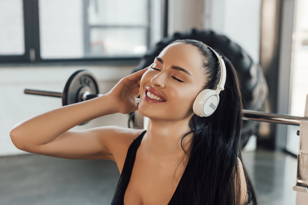 Gyönyörű sportnő csukott szemmel mosolyog és zenét hallgat a fülhallgatóban az edzőteremben - Fotó, kép