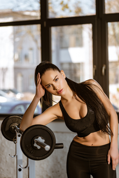 Beautiful sportswoman leaning on barbell and looking at camera in gym - Foto, afbeelding