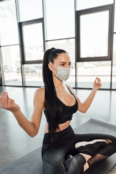 Sportnő orvosi maszkban keresztbe tett lábakkal meditál fitnesz szőnyegen a sportközpontban - Fotó, kép