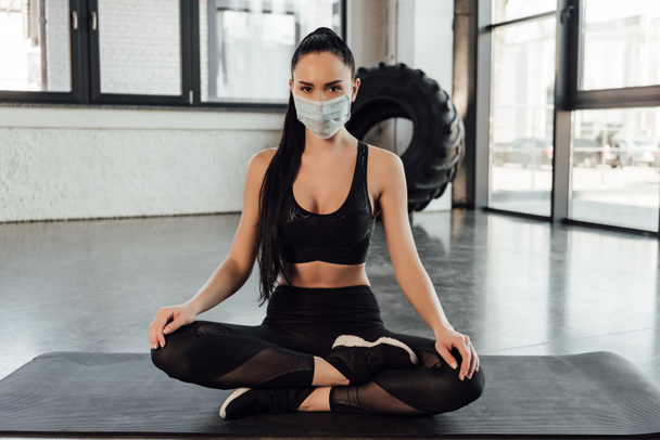 Barna sportos nő orvosi maszkban ül keresztbe tett lábakkal a fitness szőnyegen, és nézi a kamerát a tornateremben - Fotó, kép