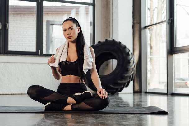 Sportswoman avec jambes croisées et serviette regardant la caméra sur tapis de fitness dans la salle de gym
 - Photo, image