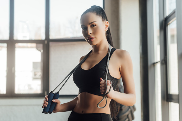 Beautiful sportswoman holding skipping rope and looking at camera in gym - Photo, Image