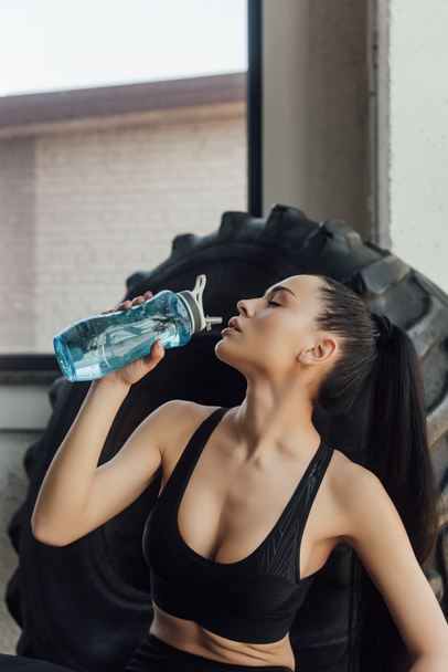 Σέξι αθλήτρια με κλειστά μάτια πίνει νερό κοντά σε λάστιχο στο γυμναστήριο - Φωτογραφία, εικόνα
